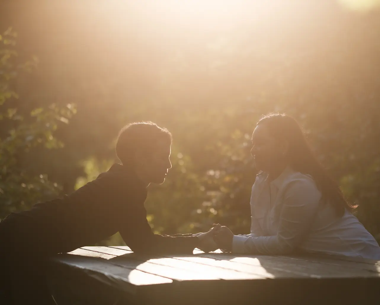 A couple leans across a picnic table, holding hands as the sun shines softly on them.